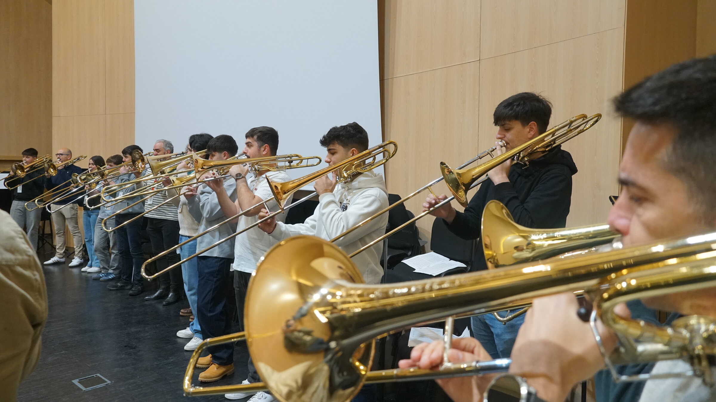 Fila de trombonistas tocando en perspectiva, cunha campá en primeiro plano
